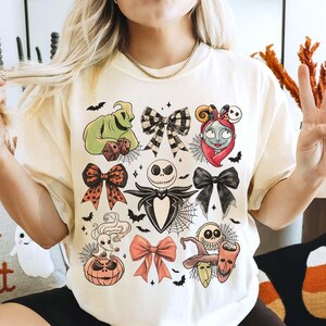 May include: Cream-coloured t-shirt featuring Halloween-themed character designs. The shirt showcases various characters and bow designs, including Jack Skellington, Sally, and Oogie Boogie. Bats and spiderwebs are also included in the design.