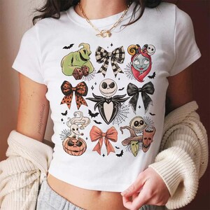 May include: White t-shirt featuring colorful illustrations of characters and bows from The Nightmare Before Christmas. The design includes Jack Skellington, Sally, Oogie Boogie, and other Halloween-themed elements.