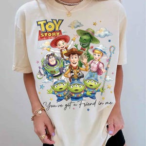 May include: Cream-colored t-shirt featuring a colorful graphic of Toy Story characters, including Woody, Buzz Lightyear, and the Little Green Men. The text "TOY STORY" and "You've got a Friend in me" are also displayed.