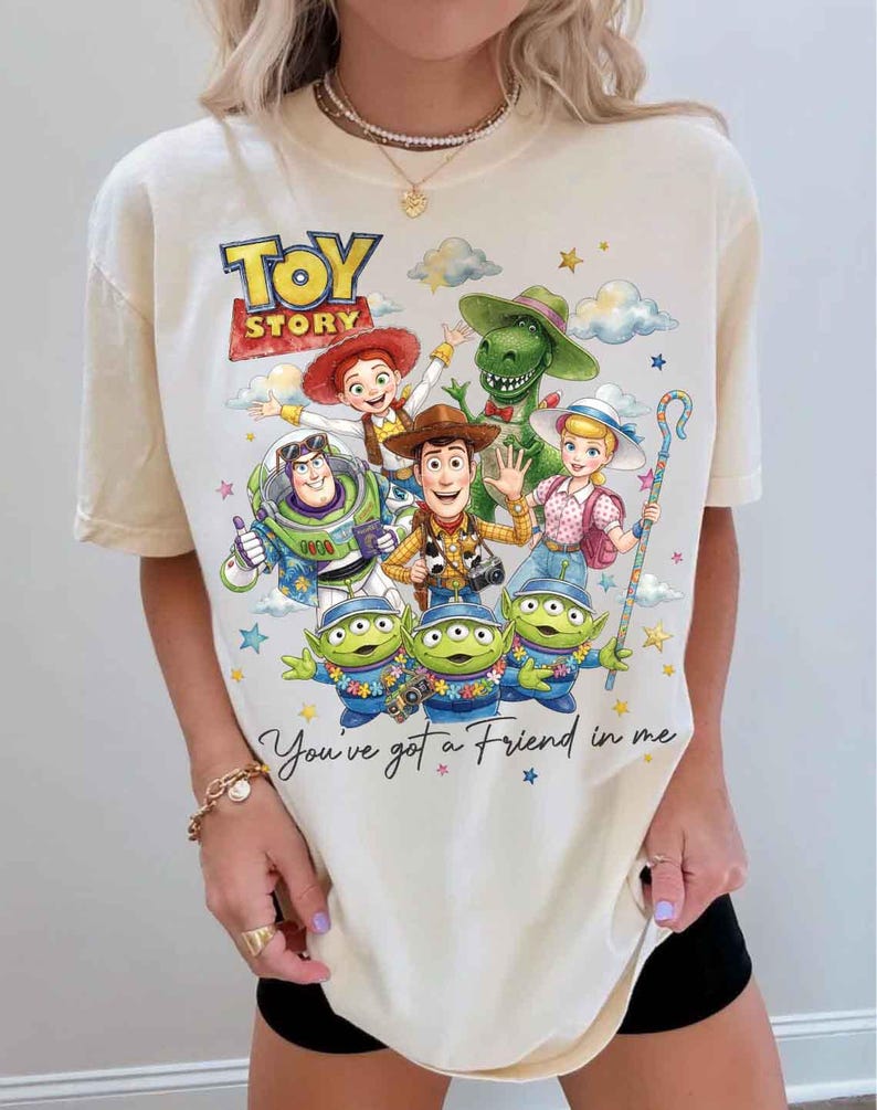May include: Cream-colored t-shirt featuring a colorful graphic of Toy Story characters, including Woody, Buzz Lightyear, and the Little Green Men. The text "TOY STORY" and "You've got a Friend in me" are also displayed.