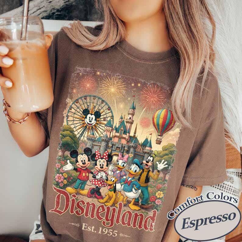 May include: A brown t-shirt featuring a graphic print of Mickey Mouse, Minnie Mouse, Donald Duck, Daisy Duck, Goofy, and Pluto in front of a castle, a Ferris wheel, and a hot air balloon with fireworks. The text reads Disney and Est. 1955.