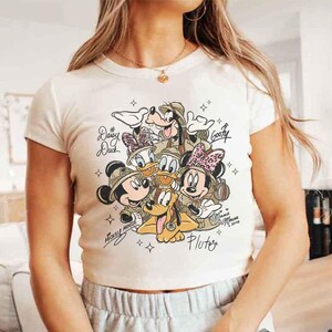 Pu&ograve; includere: T-shirt color crema a maniche corte con una grafica dei personaggi Disney Topolino, Minnie, Paperino, Paperina, Pippo e Pluto. I personaggi indossano cappelli da safari. Il testo include i nomi dei personaggi e "Minnie Mouse xoxo 2020".
