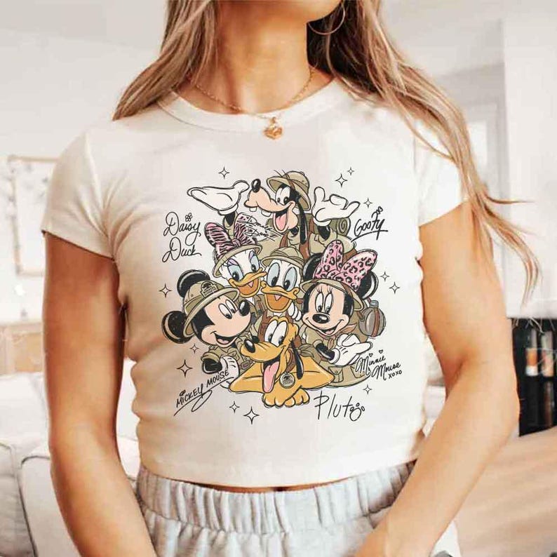 Pu&ograve; includere: T-shirt color crema a maniche corte con una grafica dei personaggi Disney Topolino, Minnie, Paperino, Paperina, Pippo e Pluto. I personaggi indossano cappelli da safari. Il testo include i nomi dei personaggi e "Minnie Mouse xoxo 2020".