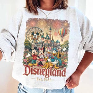 May include: A light gray crewneck sweatshirt featuring a colorful graphic of Mickey Mouse, Minnie Mouse, Pluto, Donald Duck, Daisy Duck, and Goofy in front of a castle, a Ferris wheel, and fireworks. The text reads Disneyland Est. 1955.