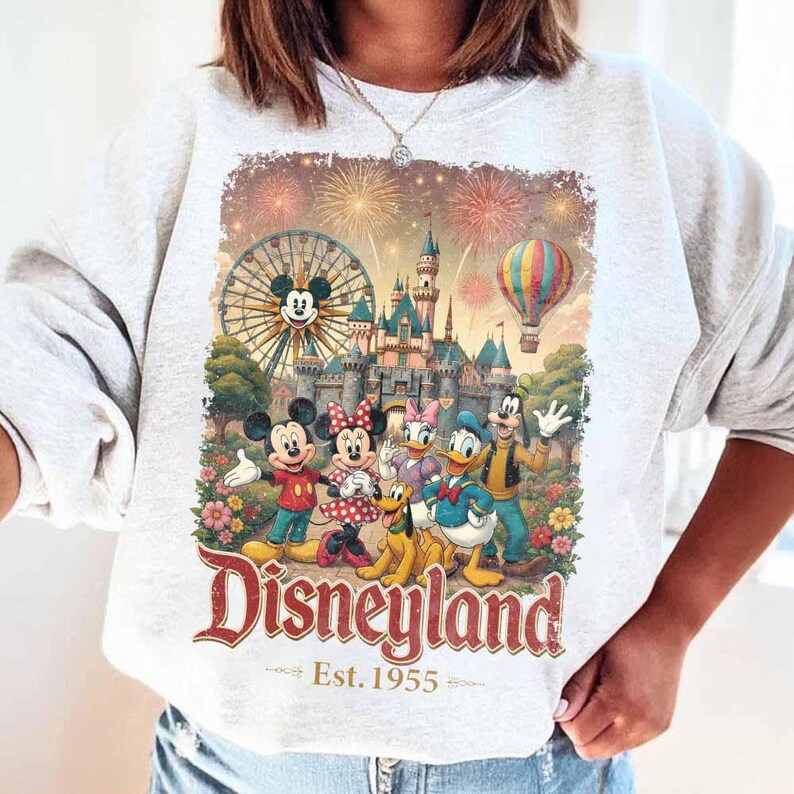 May include: A light gray crewneck sweatshirt featuring a colorful graphic of Mickey Mouse, Minnie Mouse, Pluto, Donald Duck, Daisy Duck, and Goofy in front of a castle, a Ferris wheel, and fireworks. The text reads Disneyland Est. 1955.