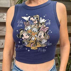 Pu&ograve; includere: Top corto blu navy con una grafica dei personaggi Disney Topolino, Minnie, Paperino, Paperina, Pippo e Pluto. I personaggi indossano cappelli da safari. Il top ha i nomi dei personaggi.