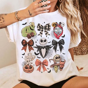 May include: White t-shirt with a colourful graphic design of characters and bows from The Nightmare Before Christmas. The design includes Jack Skellington, Sally, Oogie Boogie, and other Halloween-themed elements. Bats and spiderwebs are also included.