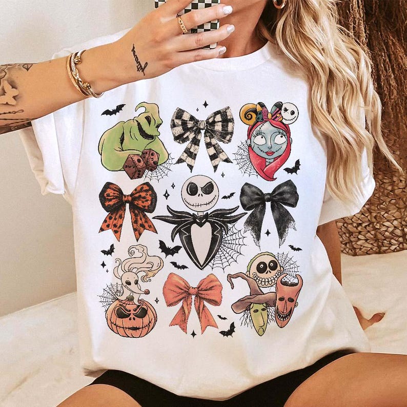 May include: White t-shirt with a colourful graphic design of characters and bows from The Nightmare Before Christmas. The design includes Jack Skellington, Sally, Oogie Boogie, and other Halloween-themed elements. Bats and spiderwebs are also included.