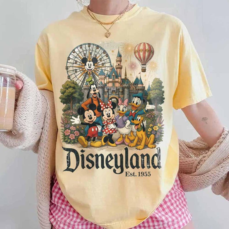May include: A pale yellow short-sleeve t-shirt featuring a graphic print of Mickey Mouse, Minnie Mouse, Goofy, Donald Duck, Daisy Duck, and Pluto in front of a castle, a ferris wheel, and a hot air balloon with the text Disneyland Est. 1955.