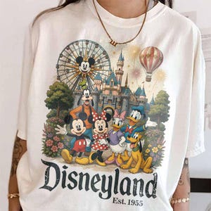 May include: A white t-shirt featuring a graphic print of Mickey Mouse, Minnie Mouse, Goofy, Donald Duck, Daisy Duck, and Pluto in front of a castle, a Ferris wheel, and a hot air balloon. The text reads Disneyland Est. 1955 in black lettering.