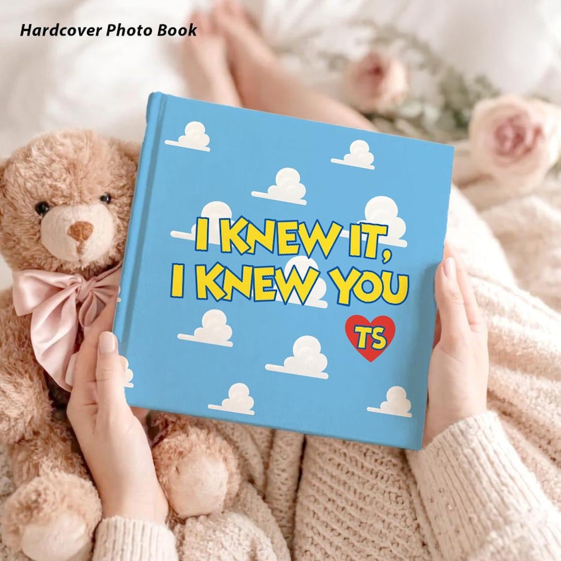 May include: A square hardcover photo book with a light blue cover featuring a white cloud pattern. The text reads I KNEW IT, I KNEW YOU in yellow letters with a red heart containing the letters TS. The book is held next to a brown teddy bear.