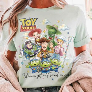 May include: Light blue t-shirt featuring a colorful graphic of Toy Story characters, including Woody, Buzz Lightyear, and the Little Green Men. The text "Toy Story" is at the top, and "You've got a Friend in me" is at the bottom.