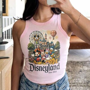 May include: A light pink sleeveless tank top featuring a graphic print of Mickey Mouse, Minnie Mouse, Goofy, Donald Duck, and Pluto in front of a castle, a Ferris wheel, and a hot air balloon. The text reads Disneyland Est. 1955 in black lettering.