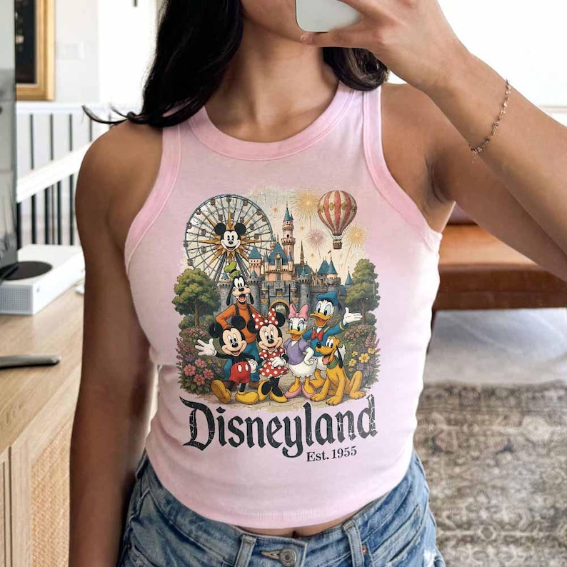 May include: A light pink sleeveless tank top featuring a graphic print of Mickey Mouse, Minnie Mouse, Goofy, Donald Duck, and Pluto in front of a castle, a Ferris wheel, and a hot air balloon. The text reads Disneyland Est. 1955 in black lettering.