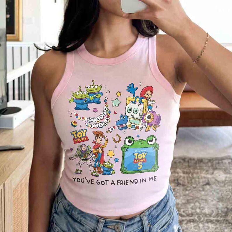 K&ouml;nnte beinhalten: Ein hellrosa &auml;rmelloses Tanktop mit einem farbenfrohen Grafikdruck von Disney Pixar Toy Story-Figuren wie Woody, Buzz Lightyear und Aliens, sowie dem aufgedruckten Text "Toy Story 5" und "You've Got A Friend In Me" auf der Vorderseite.