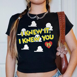 May include: A black short-sleeved t-shirt featuring a graphic print of white clouds and the yellow text "I KNEW IT, I KNEW YOU" with a red heart containing the letters "TS" in yellow. The shirt has a crew neckline and a fitted silhouette.