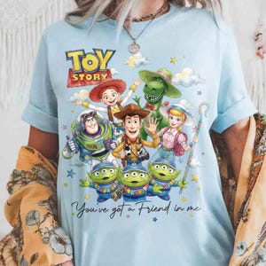 May include: Light blue t-shirt featuring a colorful graphic of Toy Story characters, including Woody, Buzz Lightyear, and the aliens. The text "TOY STORY" is at the top, with "You've got a Friend in me" below. The shirt has short sleeves.