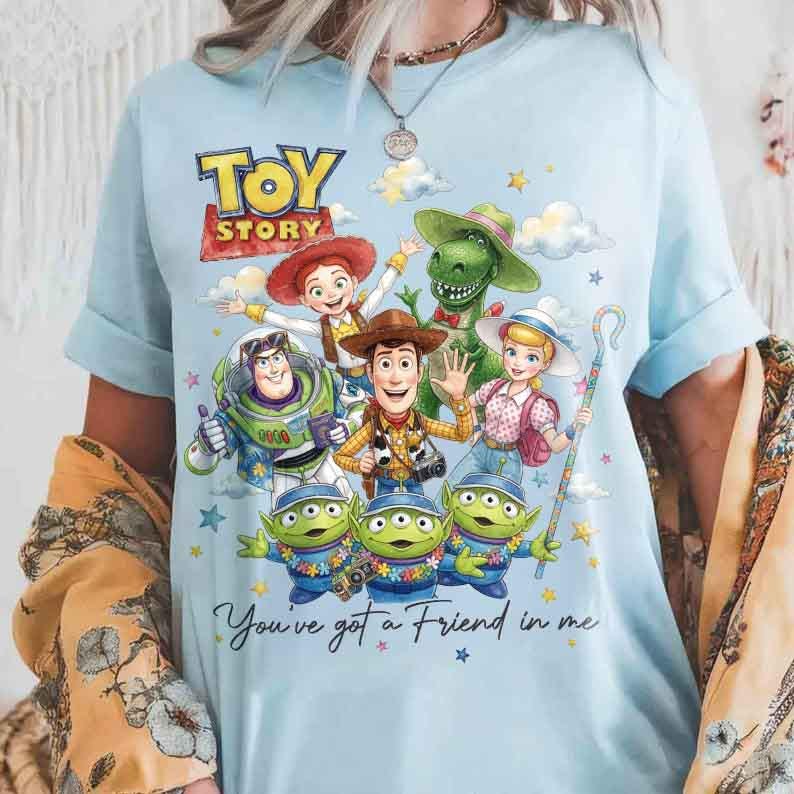 May include: Light blue t-shirt featuring a colorful graphic of Toy Story characters, including Woody, Buzz Lightyear, and the aliens. The text "TOY STORY" is at the top, with "You've got a Friend in me" below. The shirt has short sleeves.