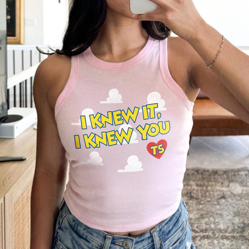 May include: A light pink sleeveless tank top with a ribbed neckline and armholes. The shirt features a repeating white cloud pattern and the yellow text "I KNEW IT, I KNEW YOU" with a blue outline, accompanied by a red heart containing the letters "TS".