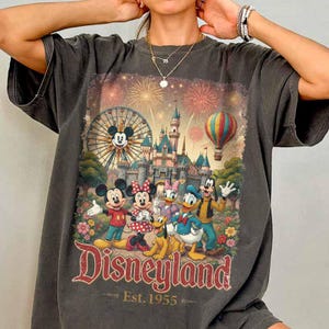 May include: A charcoal gray t-shirt featuring a graphic print of Mickey Mouse, Minnie Mouse, Donald Duck, Daisy Duck, Goofy, and Pluto in front of a castle, a Ferris wheel, and a hot air balloon with fireworks. The text reads Disneyland Est. 1955.