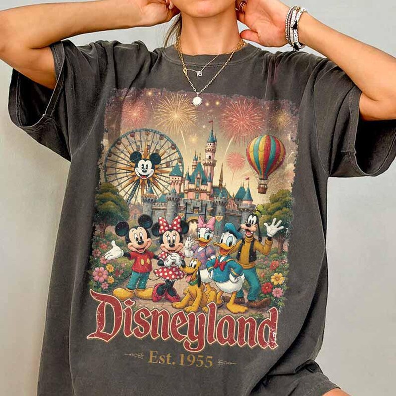 May include: A charcoal gray t-shirt featuring a graphic print of Mickey Mouse, Minnie Mouse, Donald Duck, Daisy Duck, Goofy, and Pluto in front of a castle, a Ferris wheel, and a hot air balloon with fireworks. The text reads Disneyland Est. 1955.