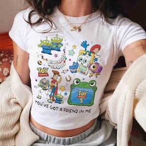 K&ouml;nnte beinhalten: Ein wei&szlig;es Kurzarm-T-Shirt mit einem farbenfrohen Grafikdruck von Toy Story-Figuren, darunter Woody, Buzz Lightyear und Aliens. Das Design enth&auml;lt den Text Disney Pixar, Toy Story, Toy Story 5 und You've Got A Friend In Me.
