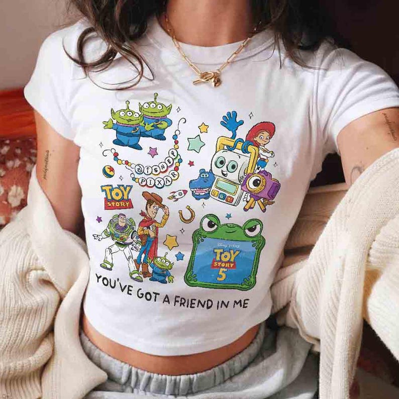 K&ouml;nnte beinhalten: Ein wei&szlig;es Kurzarm-T-Shirt mit einem farbenfrohen Grafikdruck von Toy Story-Figuren, darunter Woody, Buzz Lightyear und Aliens. Das Design enth&auml;lt den Text Disney Pixar, Toy Story, Toy Story 5 und You've Got A Friend In Me.