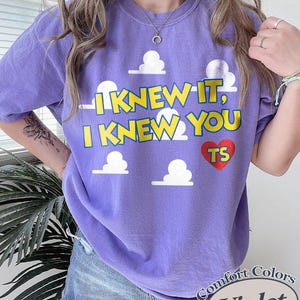 Może przedstawiać: Fioletowy t-shirt z kr&oacute;tkim rękawem z nadrukiem białych chmur i ż&oacute;łtym napisem "I KNEW IT, I KNEW YOU" oraz czerwonym sercem z literami "TS". Koszulka ma okrągły dekolt i luźny, swobodny kr&oacute;j, kt&oacute;ry zapewnia wygodę podczas codziennego noszenia.