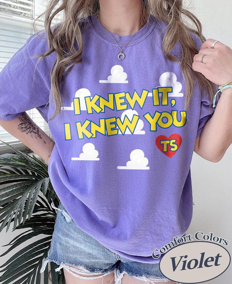 Może przedstawiać: Fioletowy t-shirt z kr&oacute;tkim rękawem z nadrukiem białych chmur i ż&oacute;łtym napisem "I KNEW IT, I KNEW YOU" oraz czerwonym sercem z literami "TS". Koszulka ma okrągły dekolt i luźny, swobodny kr&oacute;j, kt&oacute;ry zapewnia wygodę podczas codziennego noszenia.