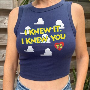 May include: A navy blue sleeveless crop top featuring a graphic print of white clouds and the yellow text "I KNEW IT, I KNEW YOU" with a red heart containing the letters "TS" positioned below the text on the front of the garment.