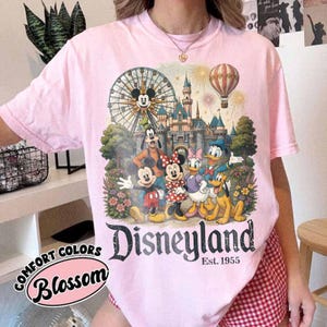May include: A light pink t-shirt featuring a graphic print of Mickey Mouse, Minnie Mouse, Goofy, Donald Duck, Daisy Duck, and Pluto in front of a castle, a Ferris wheel, and a hot air balloon. The text Disneyland Est. 1955 is printed below.
