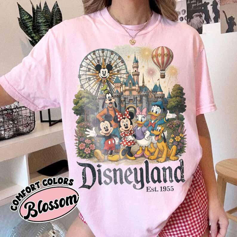 May include: A light pink t-shirt featuring a graphic print of Mickey Mouse, Minnie Mouse, Goofy, Donald Duck, Daisy Duck, and Pluto in front of a castle, a Ferris wheel, and a hot air balloon. The text Disneyland Est. 1955 is printed below.