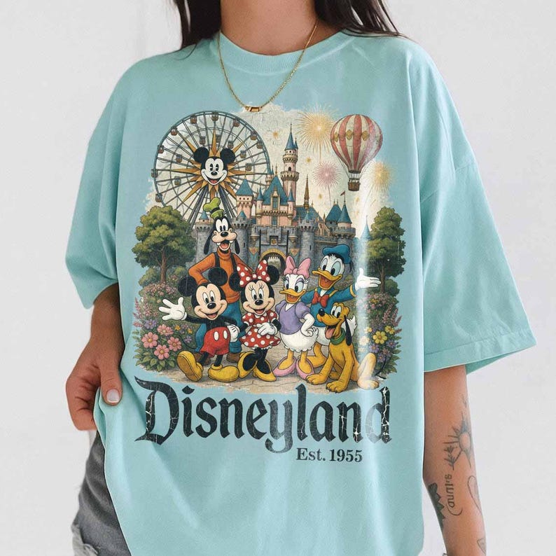 May include: A light blue t-shirt featuring a graphic print of Mickey Mouse, Minnie Mouse, Goofy, Donald Duck, Daisy Duck, and Pluto in front of a castle, a Ferris wheel, and a hot air balloon. The text Disneyland Est. 1955 is printed below.