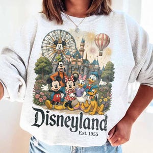 May include: A light grey crewneck sweatshirt featuring a colourful graphic of Mickey Mouse, Minnie Mouse, Goofy, Donald Duck, Daisy Duck, and Pluto in front of a castle, a Ferris wheel, and a hot air balloon. The text reads Disneyland Est. 1955.