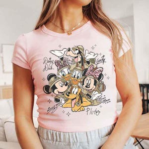 Pu&ograve; includere: T-shirt rosa chiaro con una grafica dei personaggi Disney Topolino, Minnie, Paperino, Paperina, Pippo e Pluto. I personaggi indossano cappelli da safari. Il testo include i nomi dei personaggi e "Minnie Mouse 2020".