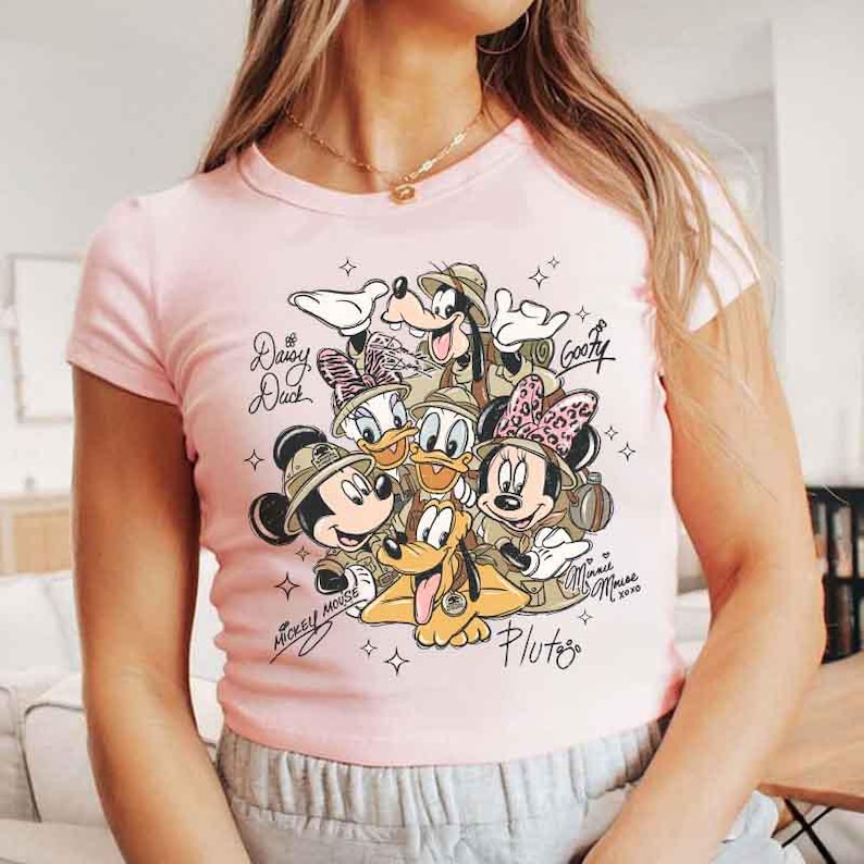 Pu&ograve; includere: T-shirt rosa chiaro con una grafica dei personaggi Disney Topolino, Minnie, Paperino, Paperina, Pippo e Pluto. I personaggi indossano cappelli da safari. Il testo include i nomi dei personaggi e "Minnie Mouse 2020".