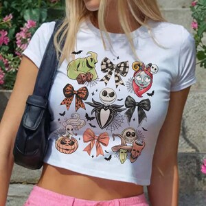 May include: White crop top featuring a colorful graphic design with characters and bows. The design includes a green Oogie Boogie, Jack Skellington, Sally, and other Halloween-themed elements. Black bats and spiderwebs are also included in the design.