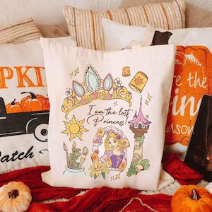 May include: Cream-colored tote bag featuring a colorful illustration of a princess, a crown, and the text "I am the lost Princess!". The design includes a sun, tower, and paintbrushes. The bag is surrounded by fall-themed pillows and pumpkins.