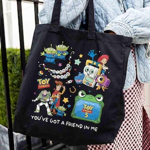 May include: A black tote bag featuring a colorful graphic print of Disney Pixar Toy Story characters including Woody, Buzz Lightyear, and aliens. The design includes the text Disney Pixar, Toy Story, Toy Story 5, and You've Got A Friend In Me.