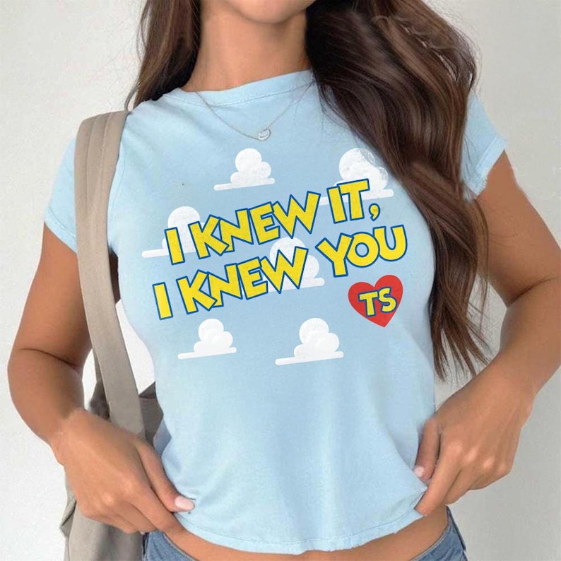 May include: A light blue short-sleeved t-shirt featuring a repeating white cloud pattern. The text "I KNEW IT, I KNEW YOU" is printed in yellow with a blue outline, accompanied by a red heart containing the letters "TS" in yellow.