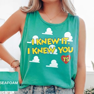 May include: A seafoam green sleeveless tank top featuring a graphic print of white clouds and the yellow text "I KNEW IT, I KNEW YOU" with a red heart containing the letters "TS". The fabric is a solid seafoam green color throughout the garment.