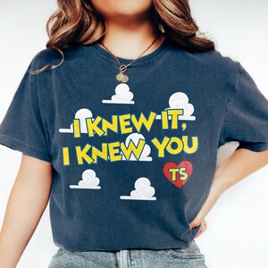 Może przedstawiać: Ciemnoniebieski t-shirt z kr&oacute;tkim rękawem, ozdobiony białymi chmurkami i ż&oacute;łtym napisem I KNEW IT, I KNEW YOU. Pod tekstem znajduje się czerwone serce z literami TS. Koszulka ma okrągły dekolt i luźny kr&oacute;j.