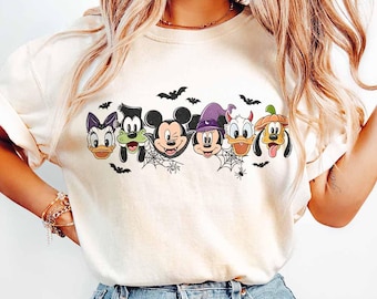 Puede incluir: Camiseta color crema con un diseño de Halloween de personajes de Disney, incluyendo Mickey Mouse, Minnie Mouse, Donald Duck y Goofy, cada uno con disfraz. Murciélagos y telarañas también están incluidos en el diseño.