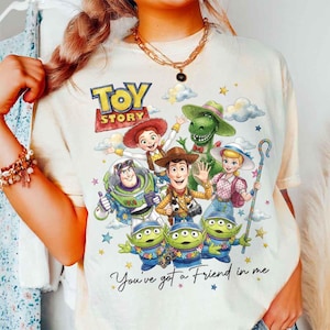 May include: Cream-colored t-shirt featuring a colorful graphic of characters from the animated film "Toy Story." The design includes Woody, Buzz Lightyear, and other characters, with the text "Toy Story" and "You've got a Friend in me."