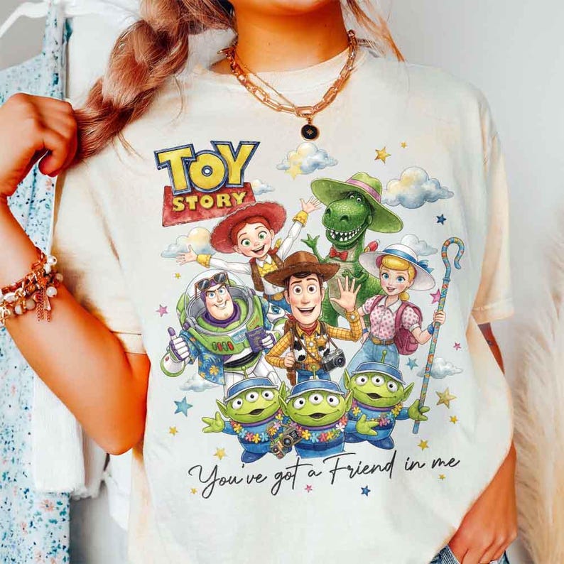 May include: Cream-colored t-shirt featuring a colorful graphic of characters from the animated film "Toy Story." The design includes Woody, Buzz Lightyear, and other characters, with the text "Toy Story" and "You've got a Friend in me."