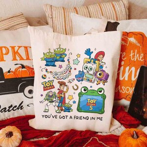 May include: A cream-colored canvas tote bag featuring a colorful graphic print of various Toy Story characters, including Woody, Buzz Lightyear, and aliens. The design includes the text "Disney Pixar," "Toy Story," "Toy Story 5," and "You've Got a Friend in Me."