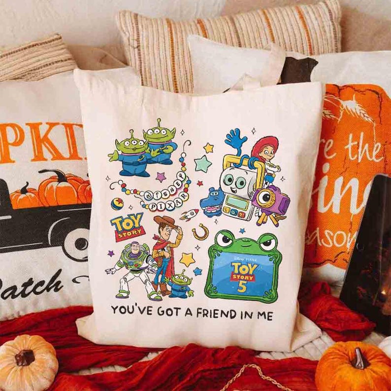 May include: A cream-colored canvas tote bag featuring a colorful graphic print of various Toy Story characters, including Woody, Buzz Lightyear, and aliens. The design includes the text "Disney Pixar," "Toy Story," "Toy Story 5," and "You've Got a Friend in Me."