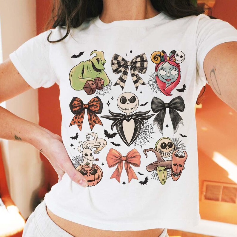 May include: White t-shirt featuring colorful illustrations of characters and bows from The Nightmare Before Christmas. The design includes Jack Skellington, Sally, Oogie Boogie, and other iconic elements, with bats and spiderwebs.
