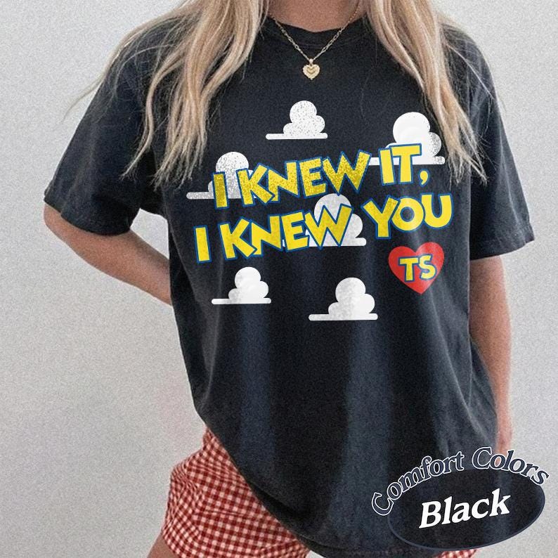 Może przedstawiać: Czarny t-shirt z kr&oacute;tkim rękawem z ż&oacute;łtym napisem "I KNEW IT, I KNEW YOU" oraz czerwonym sercem zawierającym litery "TS". Wz&oacute;r zawiera białe grafiki chmur rozmieszczone na przodzie koszulki, kt&oacute;ra ma luźny kr&oacute;j.