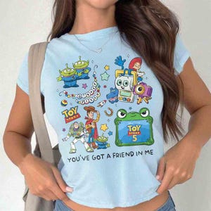 K&ouml;nnte beinhalten: Ein hellblaues T-Shirt mit einem farbenfrohen Grafikdruck der Disney Pixar Toy Story-Figuren, darunter Woody, Buzz Lightyear und gr&uuml;ne Aliens. Das Design enth&auml;lt den Text Toy Story, Toy Story 5 und You've Got a Friend in Me.